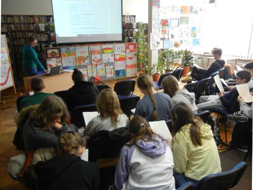 Widok od tyłu na młodzież uczestniczącą w zajęciach w bibliotece, w tle widać prowadzącą dr Barbarę Męczykowską oraz ekran z prezentacją.